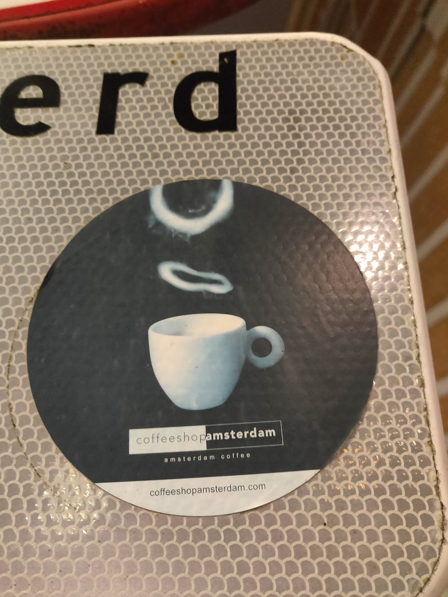 Round sticker with a picture of a cup of coffee and the text "coffeeshopamsterdam" and the website address. The design is minimalist with a dark background and a light-colored cup.