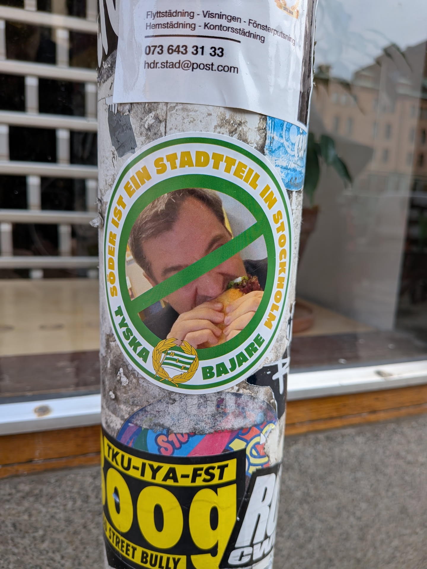 A circular sticker featuring a man eating a burger, overlaid with a green 'no' symbol (a circle with a diagonal line). The man has light brown hair and is seen from the chest up, with only part of his face visible behind the burger. The outer ring of the sticker is green, with yellow text on the top half reading 'SÖDER IST EIN STADTTEIL IN STOCKHOLM' and green text on the bottom half reading 'TYKSA BAJARE'. At the very bottom, centered, is a small emblem resembling a flag or crest with green and white stripes, possibly the Hammarby IF (HIF) logo, surrounded by a golden wreath. The sticker has a white inner border separating the central image from the text ring.