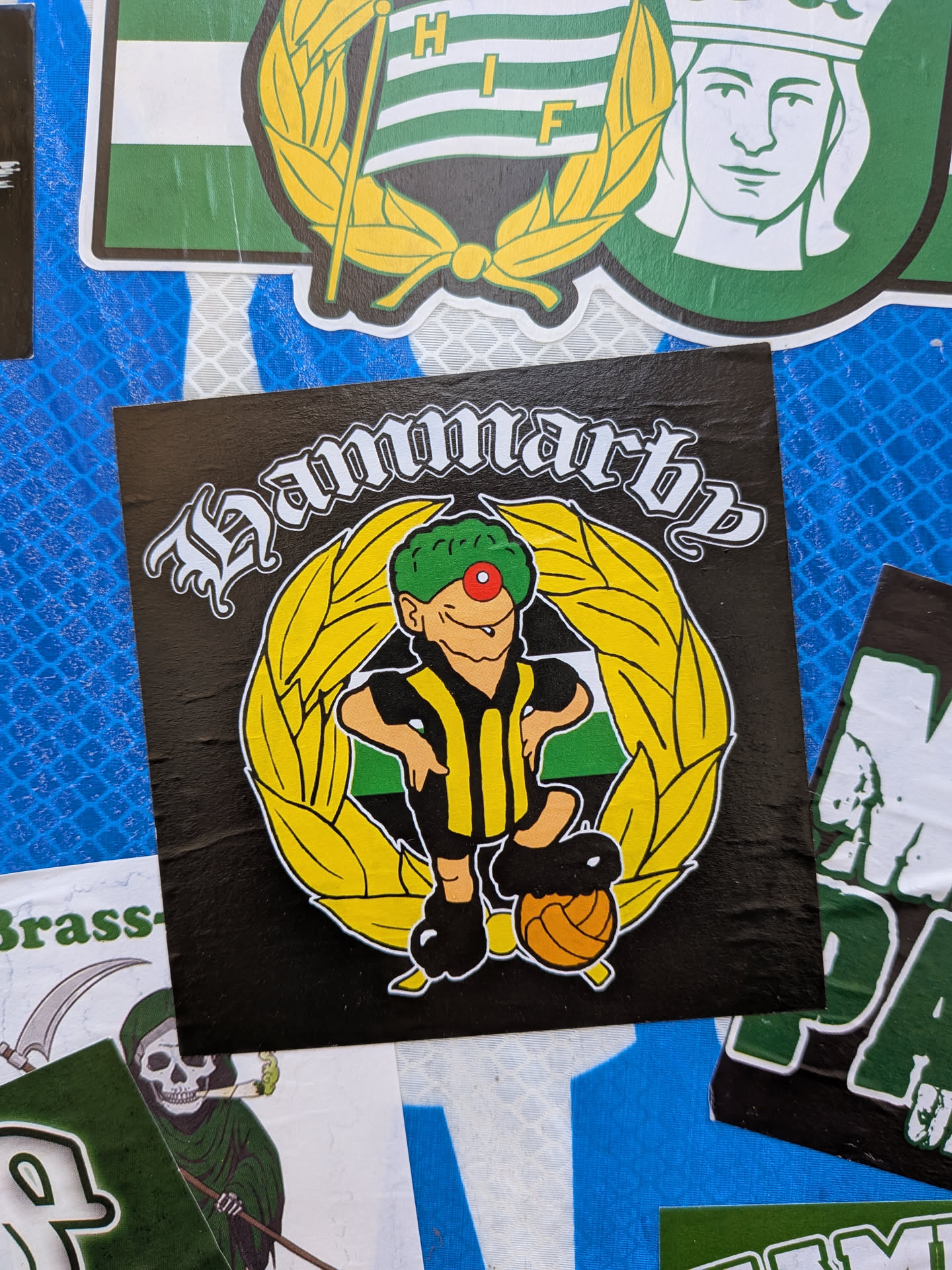 A square black sticker featuring a cartoon mascot character. The character has green hair, a red clown nose, and a wide smile, wearing a yellow and black striped shirt, black shorts, and black shoes. He stands with hands on hips, with a brown/orange football at his left foot. He is encircled by a prominent yellow laurel wreath. Behind the character, within the wreath, is a green and white striped shield or flag. Above the wreath, the word 'Hammarby' is written in a distinctive white gothic-style font. The overall design is vibrant with black, yellow, green, white, and red colors.