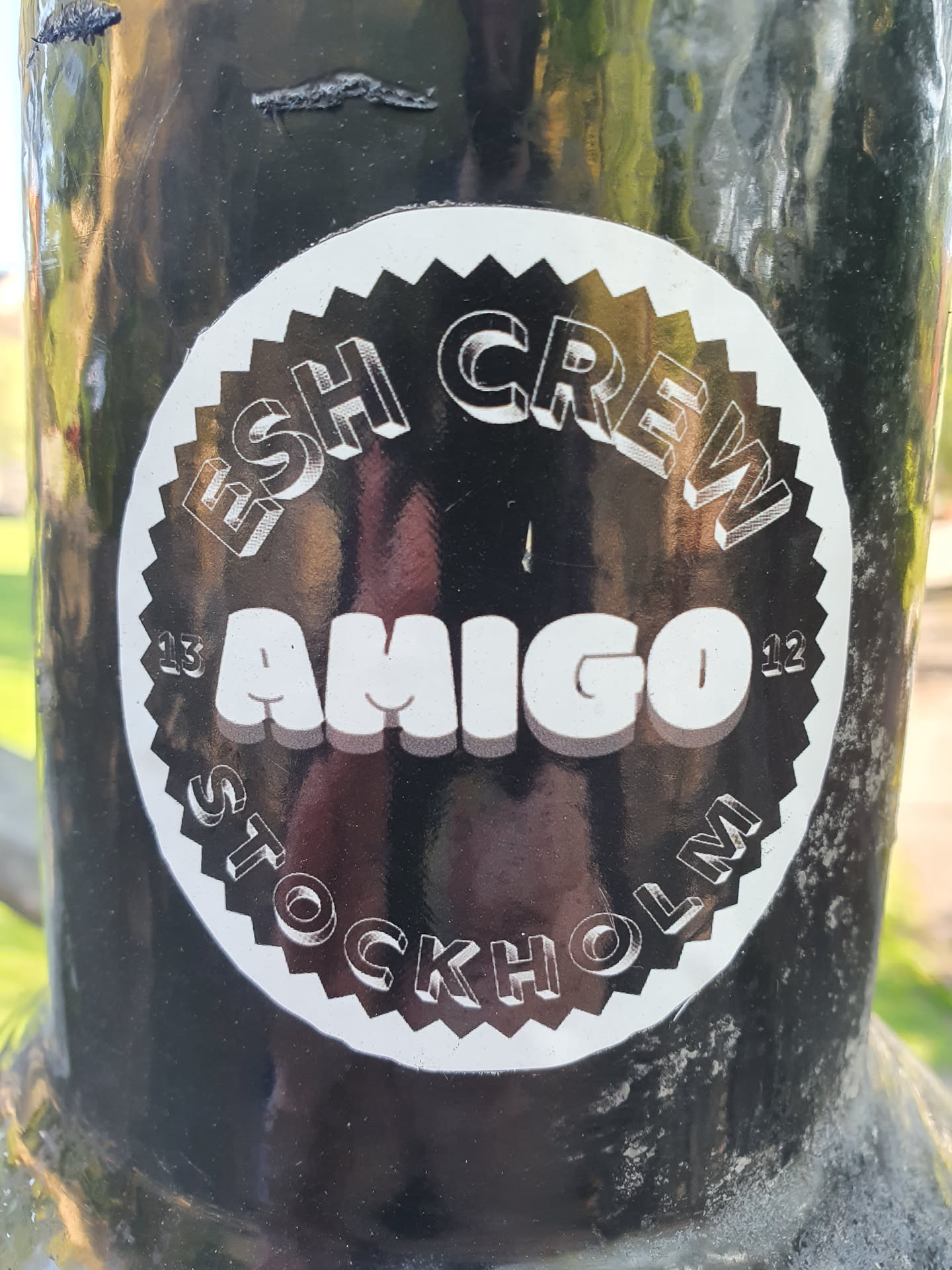 Black and white circular sticker with the text "ESH CREW" at the top, "AMIGO" in the center, and "STOCKHOLM" at the bottom. The text has a 3D effect. The sticker is on a dark surface.