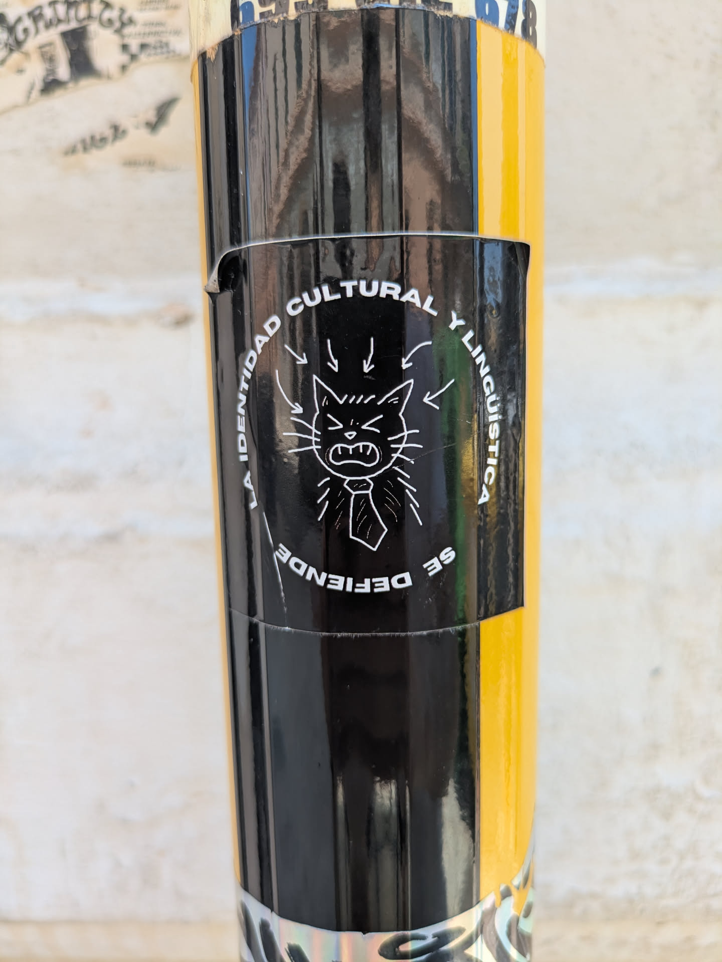 The black and white sticker features a drawing of an angry cat wearing a tie. The text surrounding the cat reads "LA IDENTIDAD CULTURAL Y LINGUISTICA SE DEFIENDE".