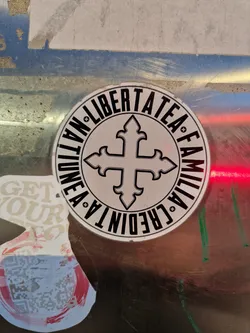 A round sticker with the text "NATION", "LIBERTATEA", "FAMILIA", "CREDO" encircling a cross symbol. The text is in a circular pattern around the cross.