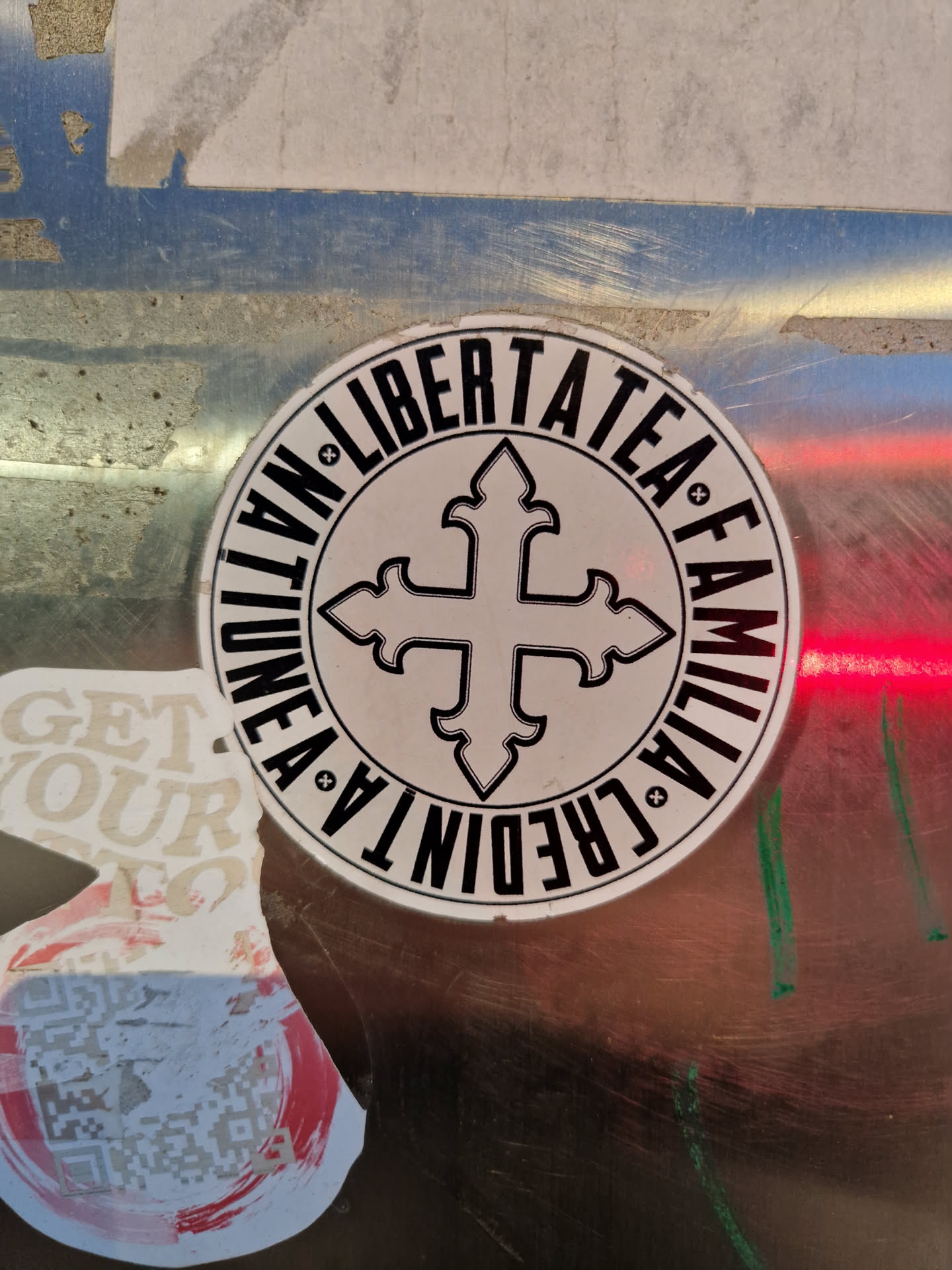 A round sticker with the text "NATION", "LIBERTATEA", "FAMILIA", "CREDO" encircling a cross symbol. The text is in a circular pattern around the cross.