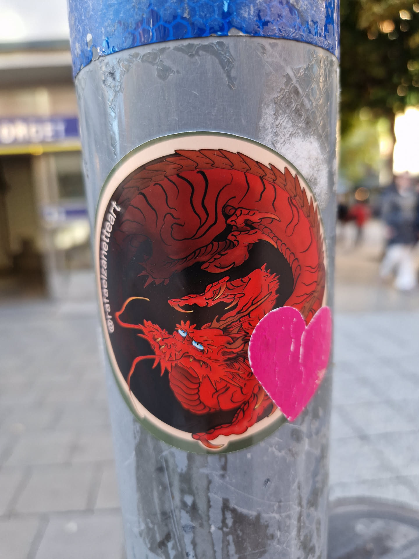A round sticker featuring a detailed illustration of a red dragon. A smaller pink heart sticker is adhered to the side of the main sticker. The dragon is depicted in a circular design, and the style suggests Asian influences. The sticker is affixed to a gray metal pole outdoors.