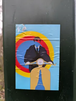 The sticker features a man wearing a black suit and tie, only his silhouette is visible, against a target background of red, yellow, and blue circles. He is riding a yellow scooter. The sticker is affixed to a green pole.