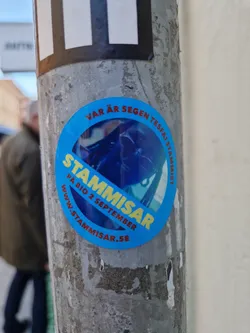 Round blue sticker with the text "Stämmisar" in large yellow letters.  Smaller text says "På Bio 2 September" and a website address: www.stammisar.se. There is also some smaller text in a circle around the edge which is partially obscured, but seems to contain the words "Vår är", and the word "Stämmisar" repeated. The central image is a blurry photo of what looks like a person or creature possibly wearing a helmet, in a dark blue tone. 