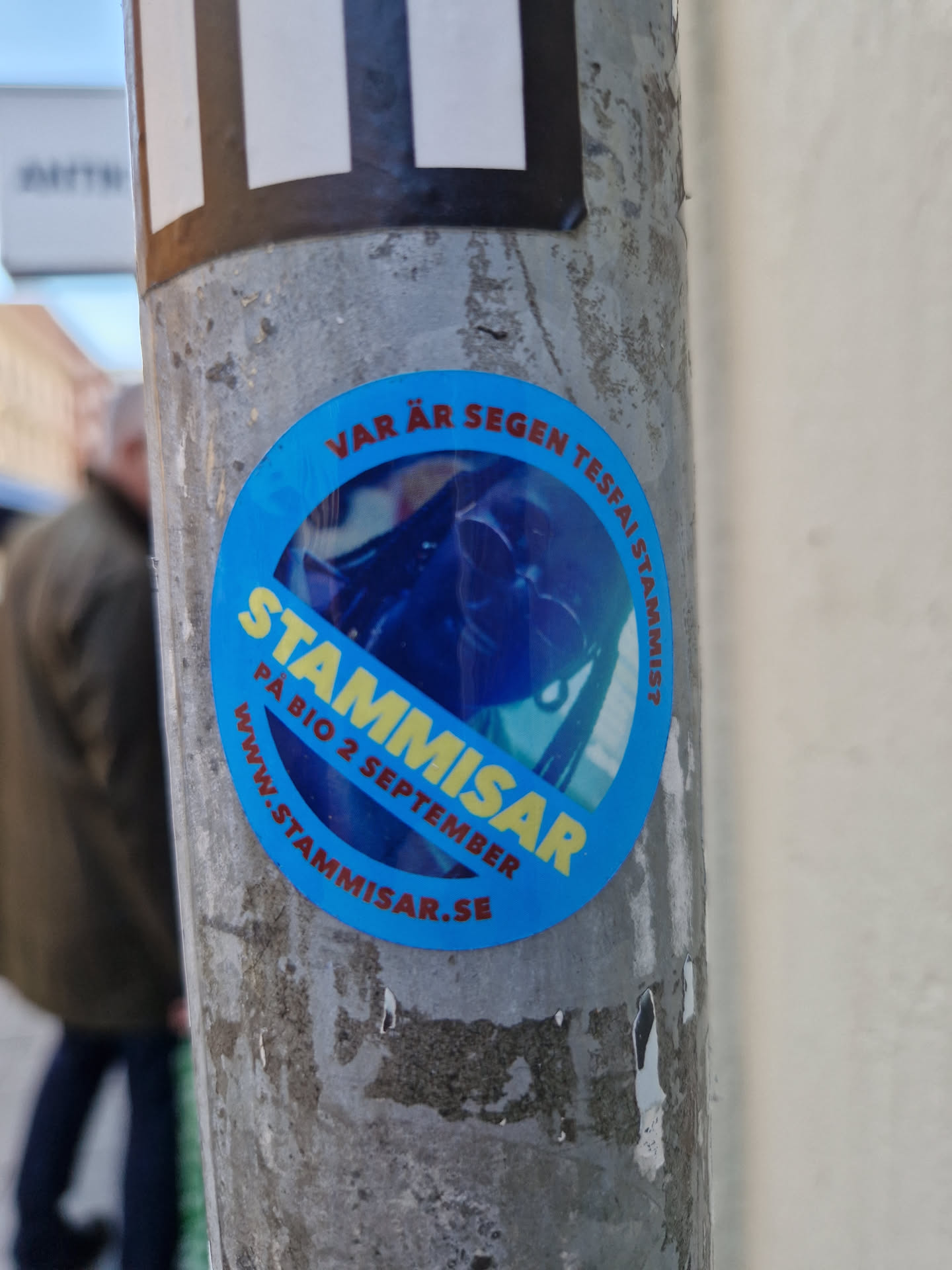 Round blue sticker with the text "Stämmisar" in large yellow letters.  Smaller text says "På Bio 2 September" and a website address: www.stammisar.se. There is also some smaller text in a circle around the edge which is partially obscured, but seems to contain the words "Vår är", and the word "Stämmisar" repeated. The central image is a blurry photo of what looks like a person or creature possibly wearing a helmet, in a dark blue tone. 