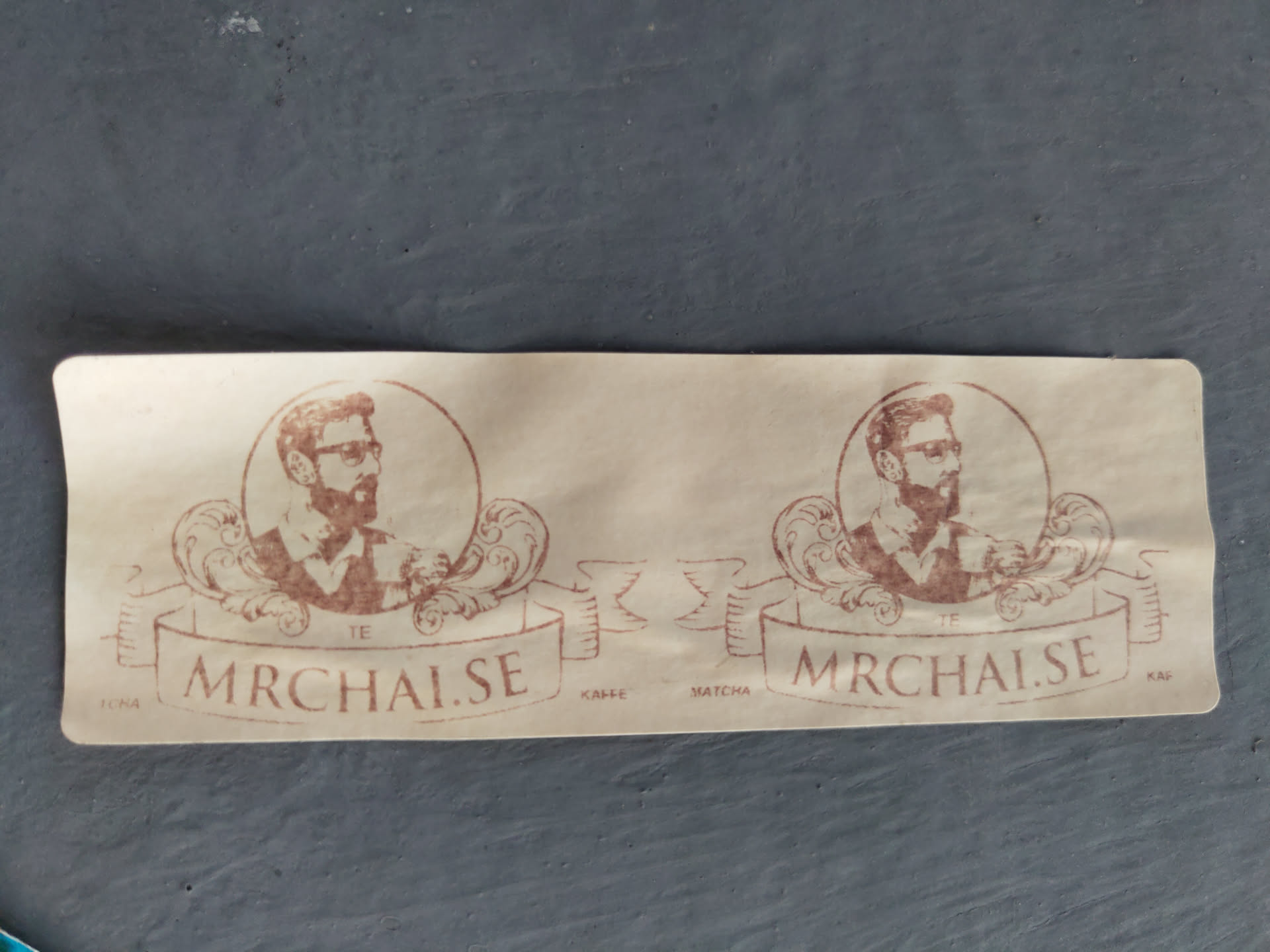 A beige sticker featuring a repeating design. Each element includes a portrait of a man with a beard and glasses set within a circular frame, accented by ornate flourishes. A banner beneath reads "mrchai.se". The overall aesthetic is vintage or retro.