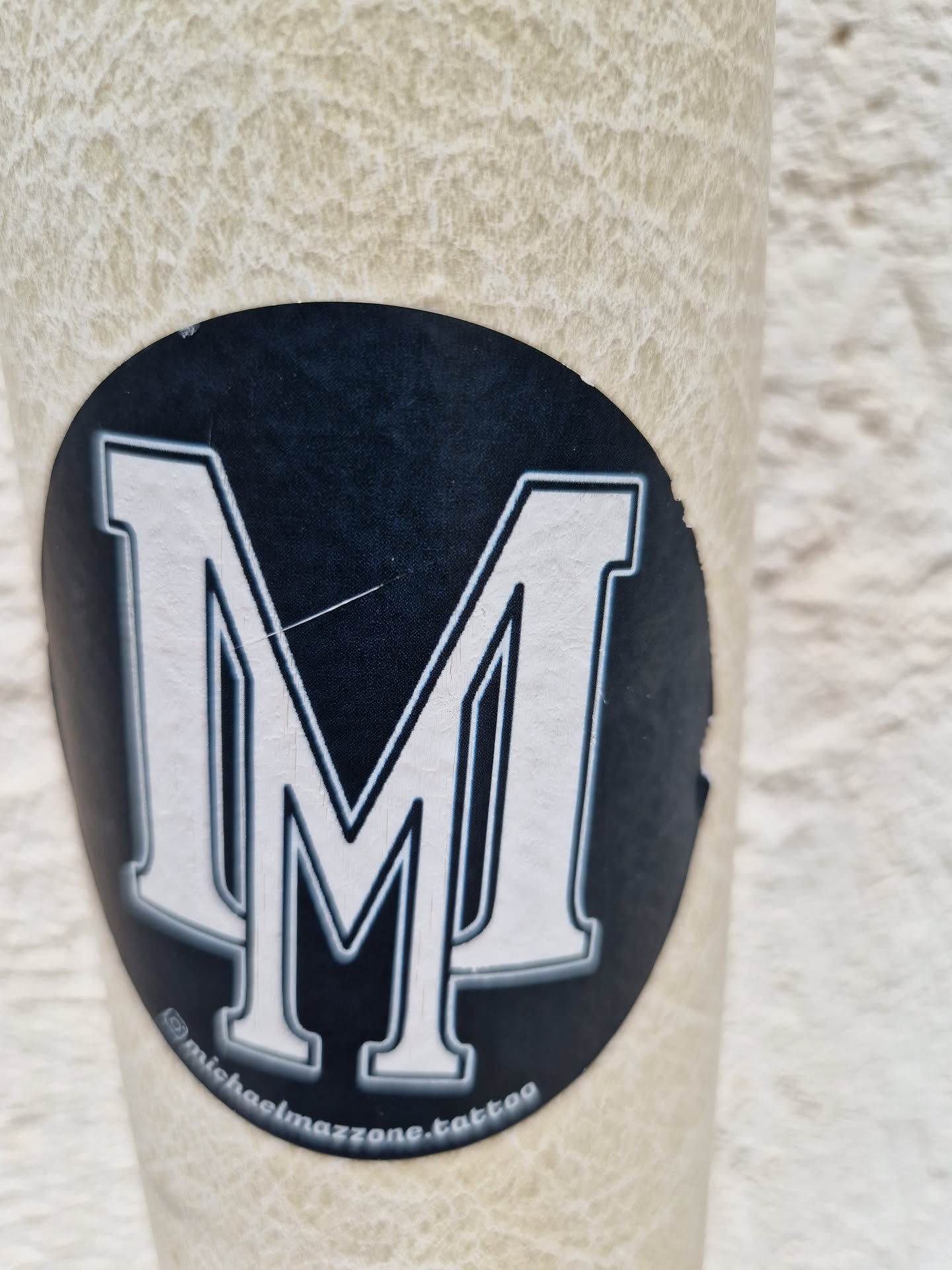 This sticker features a bold, uppercase letter "M" in a classic, serif typeface.  The "M" is white with a black outline, set against a black circular background.  Small text at the bottom reads "@michaelmazzone.tattoo".