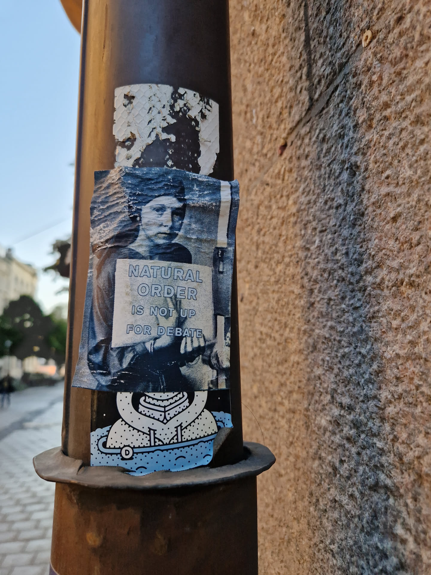 A sticker featuring a black and white image of a person holding a sign that says "Natural Order Is Not Up For Debate". Below it, there's a smaller sticker of a stylized figure in a spacesuit. The stickers are attached to a metal pole.