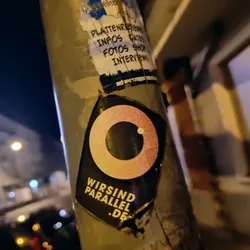 A black and peach circular sticker with the text 'wirSind parallel .de' at the bottom.  The main graphic is an eye-like symbol.