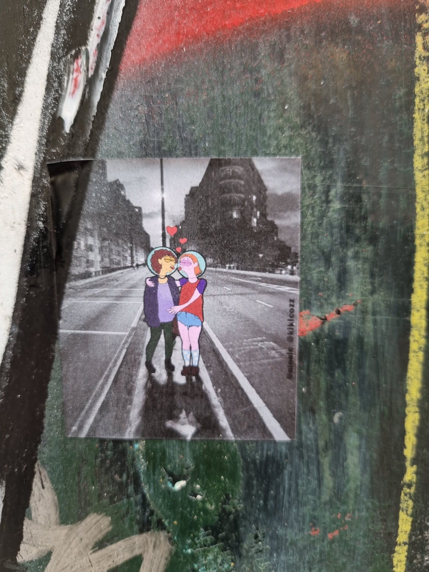 This sticker features a black and white illustration of two girls sharing a kiss in the middle of a city street. The girls are depicted in a simple, almost childlike style, while the background features realistic buildings and a street scene. It is a very cute and romantic image.