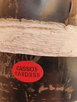A close-up shot of a red oval sticker affixed to a dark-colored surface. The sticker features the text "((ASSO))" stacked above "((A)(D)(S))" in a sans-serif font. The background shows signs of wear and tear, possibly indicating the sticker has been placed outdoors for a period of time. The overall impression is slightly gritty and urban.