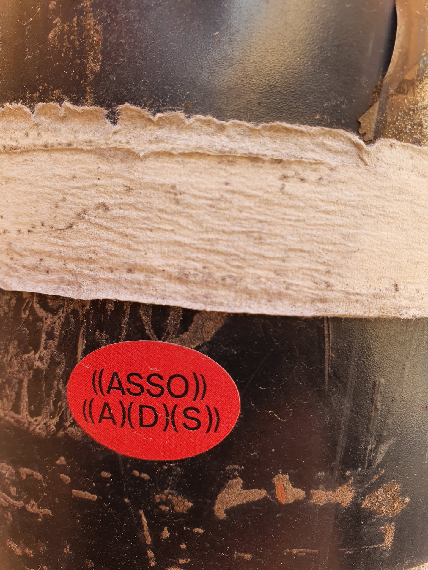 A close-up shot of a red oval sticker affixed to a dark-colored surface. The sticker features the text "((ASSO))" stacked above "((A)(D)(S))" in a sans-serif font. The background shows signs of wear and tear, possibly indicating the sticker has been placed outdoors for a period of time. The overall impression is slightly gritty and urban.