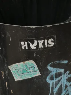 The sticker is a rectangular, monochrome design featuring white elements on a black background. It displays the letters 'H' and 'KIS' in a bold, sans-serif font. Between the 'H' and 'KIS', a stylized white silhouette of an eagle in flight is prominently featured, appearing to replace or represent a letter within the word. The eagle is depicted with outstretched wings, in a dynamic pose. The sticker shows signs of wear, with some peeling and minor damage, particularly around its edges and on the white lettering, indicating it has been affixed for some time on a dark, textured surface.