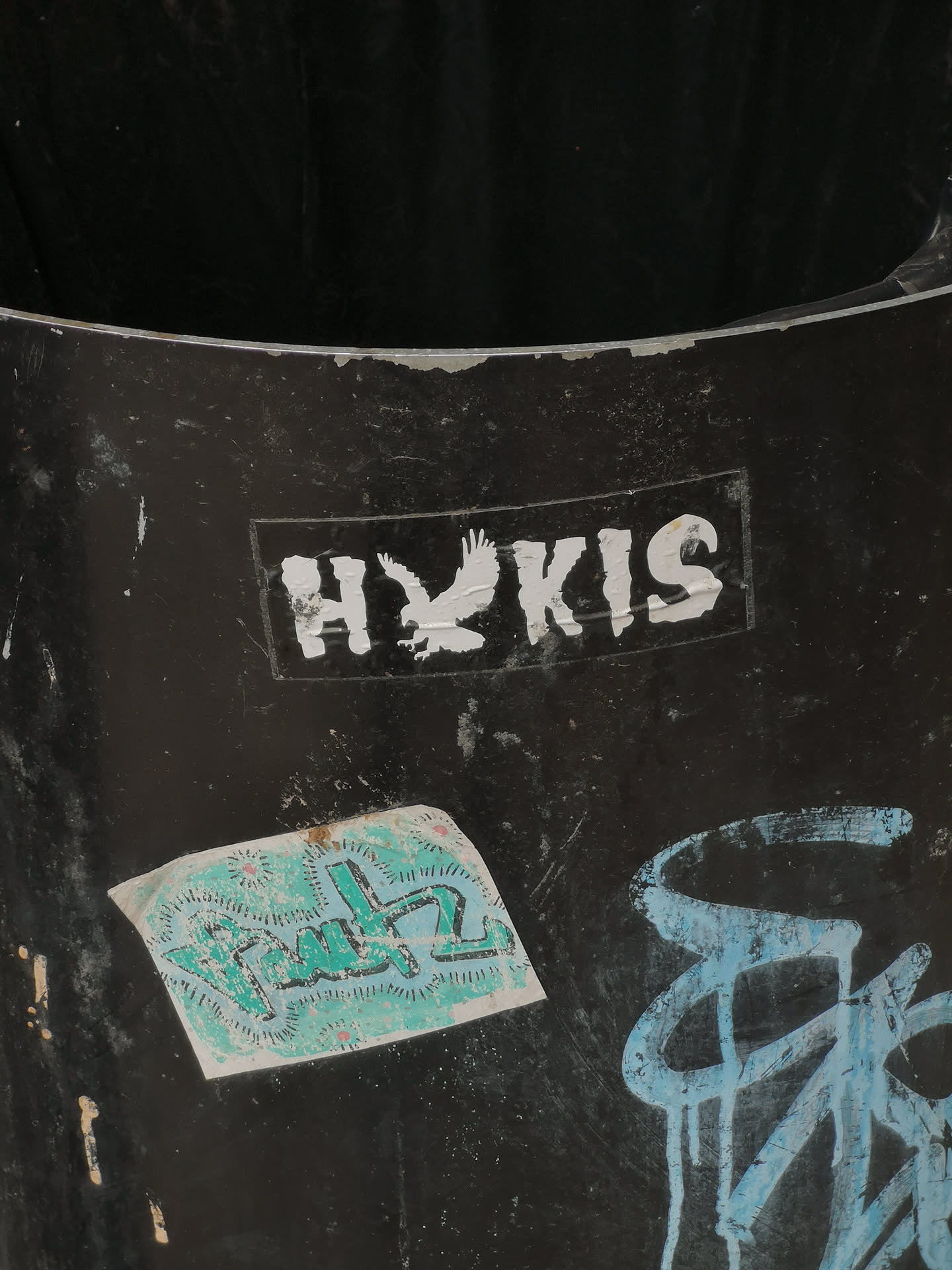 The sticker is a rectangular, monochrome design featuring white elements on a black background. It displays the letters 'H' and 'KIS' in a bold, sans-serif font. Between the 'H' and 'KIS', a stylized white silhouette of an eagle in flight is prominently featured, appearing to replace or represent a letter within the word. The eagle is depicted with outstretched wings, in a dynamic pose. The sticker shows signs of wear, with some peeling and minor damage, particularly around its edges and on the white lettering, indicating it has been affixed for some time on a dark, textured surface.
