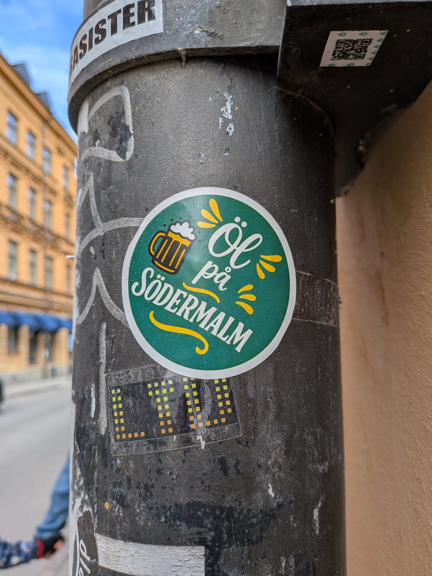 A circular sticker with a dark teal-green background and a white border. On the left, a stylized golden beer mug with white foam is depicted. To its right, the text 'Öl på' is written in a white, elegant script font, positioned above 'SÖDERMALM' in a bold, white, sans-serif, all-caps font. Yellow stylized 'splashes' or 'rays' emanate from the text, and a yellow decorative swirl underlines 'SÖDERMALM'. The overall design is clean, cheerful, and inviting.