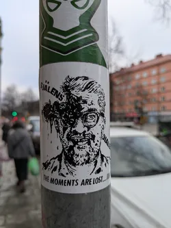 Black and white sticker featuring a stylized portrait of a man with splattered ink or blood around his head. Text in Swedish and English is visible. 