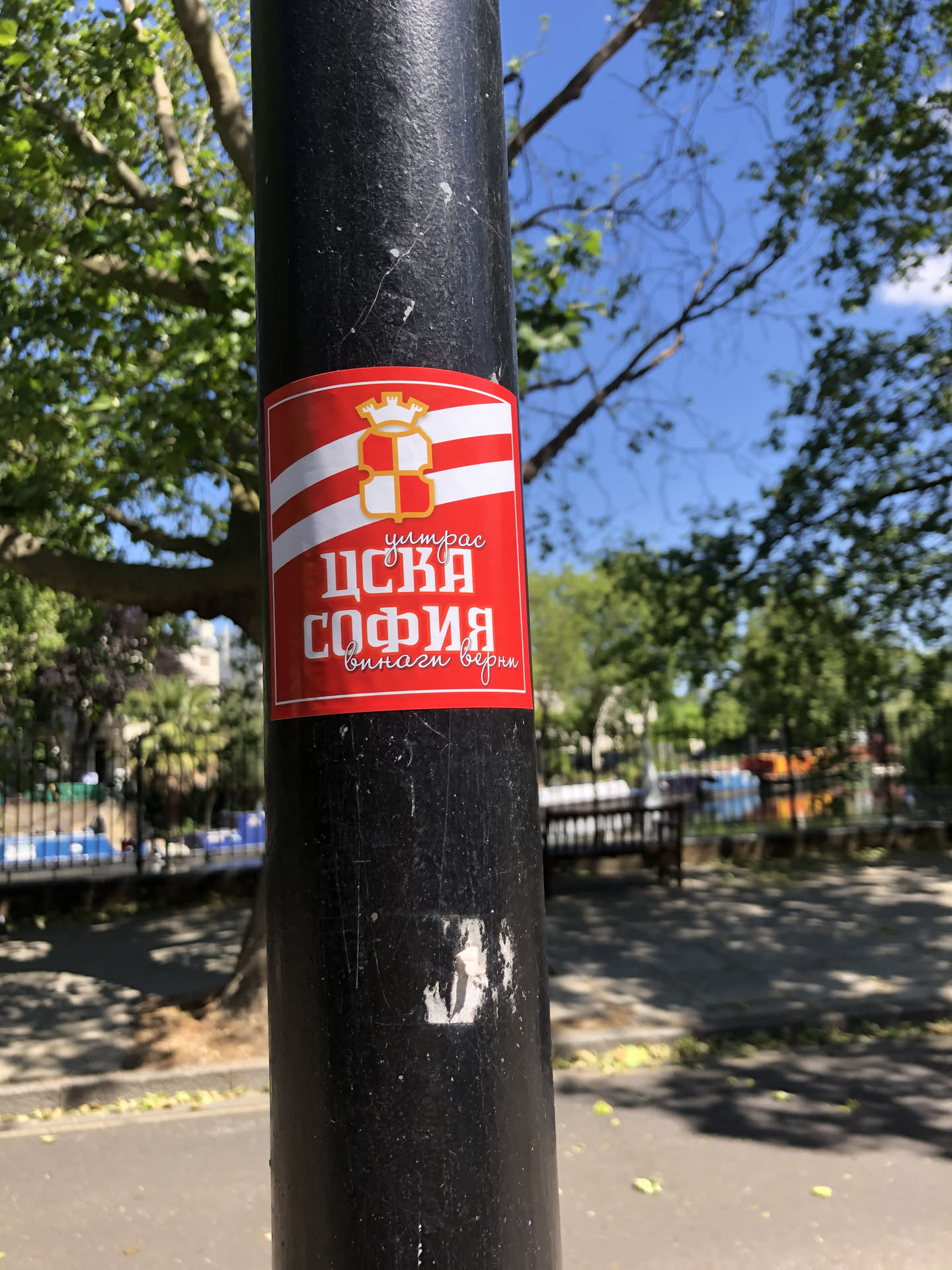 A rectangular sticker with a prominent red background, featuring a white and red striped pattern across its upper half. Centered on the stripes is a golden crest, resembling a shield with a cross design, topped by a crown or castle turret. Below the stripes, on the solid red background, is white Cyrillic text. The main text 'ЦСКА СОФИЯ' is large and bold, with smaller, cursive text 'Шитрас' above it and 'винаги верни' below. The sticker has a slight sheen and appears to be adhered to a black metal pole.