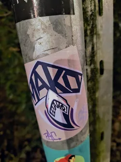 A sticker featuring a stylized mako shark with the word MAKO incorporated into the design. The color palette is primarily pink, purple, and black. A smaller signature or tag is visible near the bottom.