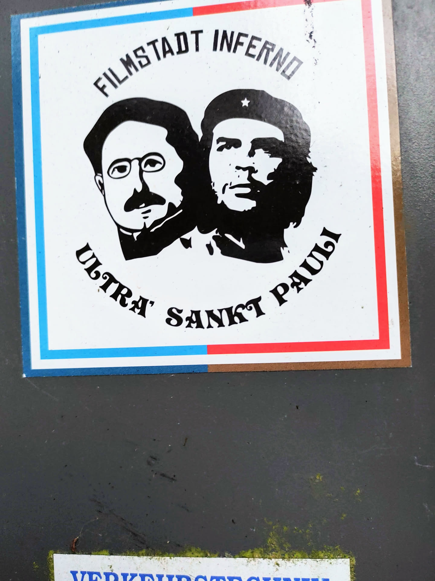A sticker featuring black and white portraits of Bertolt Brecht and Che Guevara, with text that reads 'Filmstadt Inferno Ultra Sankt Pauli'. The sticker has a tri-color design with blue, white, and red stripes.