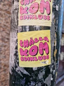 Close-up of a yellow sticker with pink lettering that says "Snälla kom ediklubb". The sticker is slightly worn and attached to a dark-colored pole.