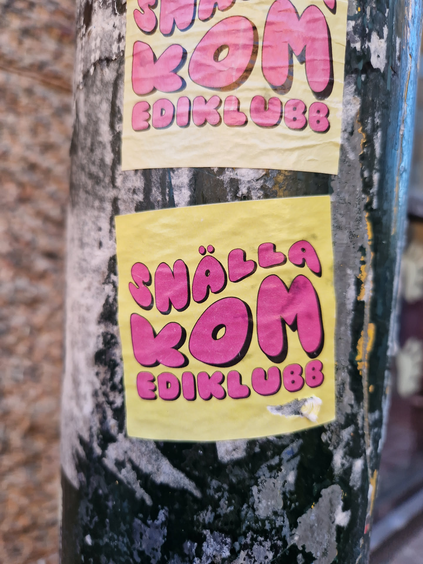 Close-up of a yellow sticker with pink lettering that says "Snälla kom ediklubb". The sticker is slightly worn and attached to a dark-colored pole.