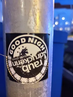 A black and white circular sticker with the text "GOOD NIGHT" at the top and "Rancid Tauckern" vertically stacked in the middle. There is an image of a unicorn with stars on top, and the text "Street Prints" is written underneath. The sticker is placed on a metallic pole.