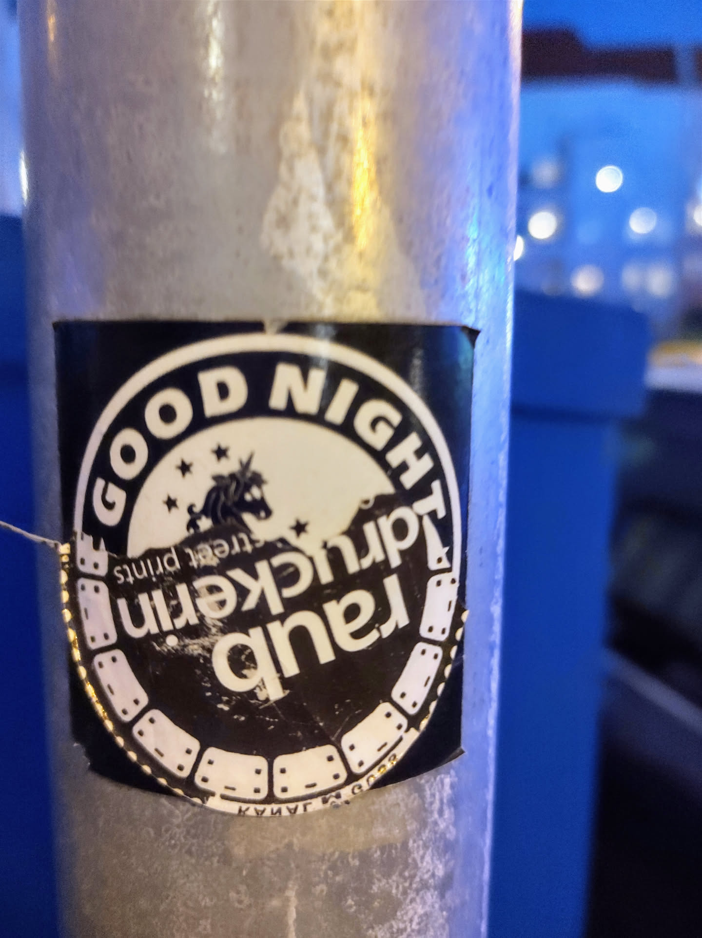 A black and white circular sticker with the text "GOOD NIGHT" at the top and "Rancid Tauckern" vertically stacked in the middle. There is an image of a unicorn with stars on top, and the text "Street Prints" is written underneath. The sticker is placed on a metallic pole.