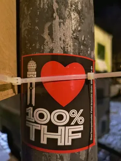 A sticker with the text "I LOVE 100% THF" on a black background with a red border. The "I" has a building with a dome on top, the word "LOVE" is replaced with a red heart and "THF" is in large white letters. The sticker is affixed to a pole using zip ties.