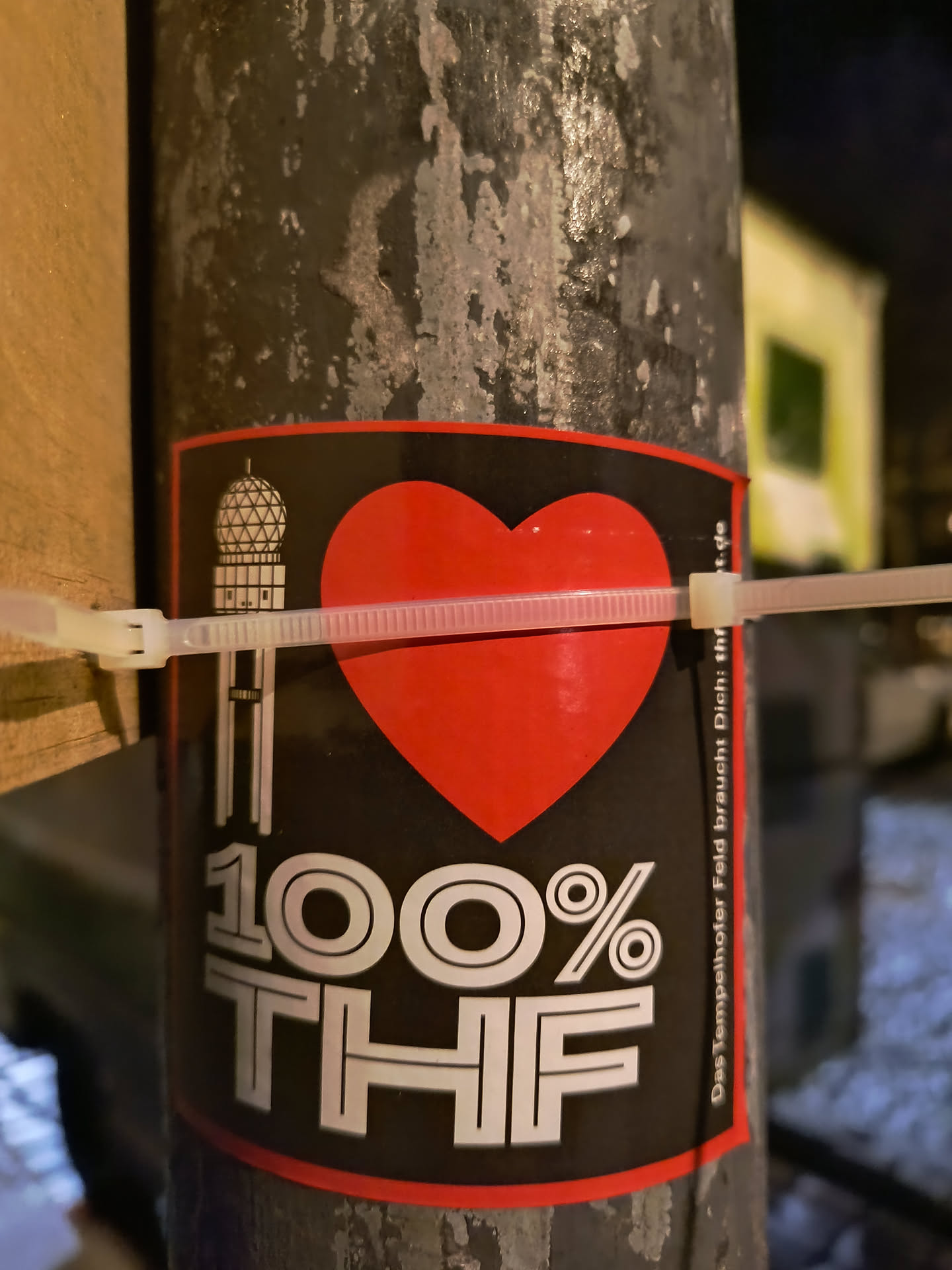 A sticker with the text "I LOVE 100% THF" on a black background with a red border. The "I" has a building with a dome on top, the word "LOVE" is replaced with a red heart and "THF" is in large white letters. The sticker is affixed to a pole using zip ties.