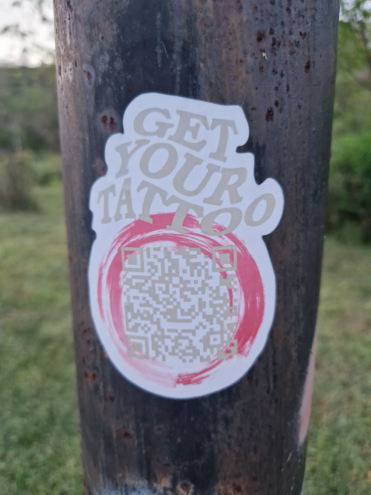 A sticker with the text "GET YOUR TATTOO" and a QR code in the center. The sticker is attached to a rusty pole outdoors. The QR code appears to link to a tattoo parlor or artist.