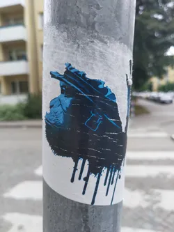 A sticker featuring a dark blue and black illustration possibly depicting a person or a creature, with a dripping paint effect at the bottom. The sticker is affixed to a gray pole and shows some wear and tear, with parts of it peeling off.