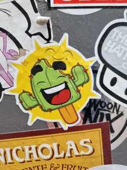 A sticker featuring a cartoon cactus popsicle with a smiling face, surrounded by yellow sun rays. The art style is playful and vibrant.