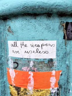 A small hand-written white sticker affixed to a teal-colored surface. The sticker is slightly weathered and torn at the edges. The text on the sticker reads, "All the weapons are useless". The background is a weathered teal surface with other stickers and markings.