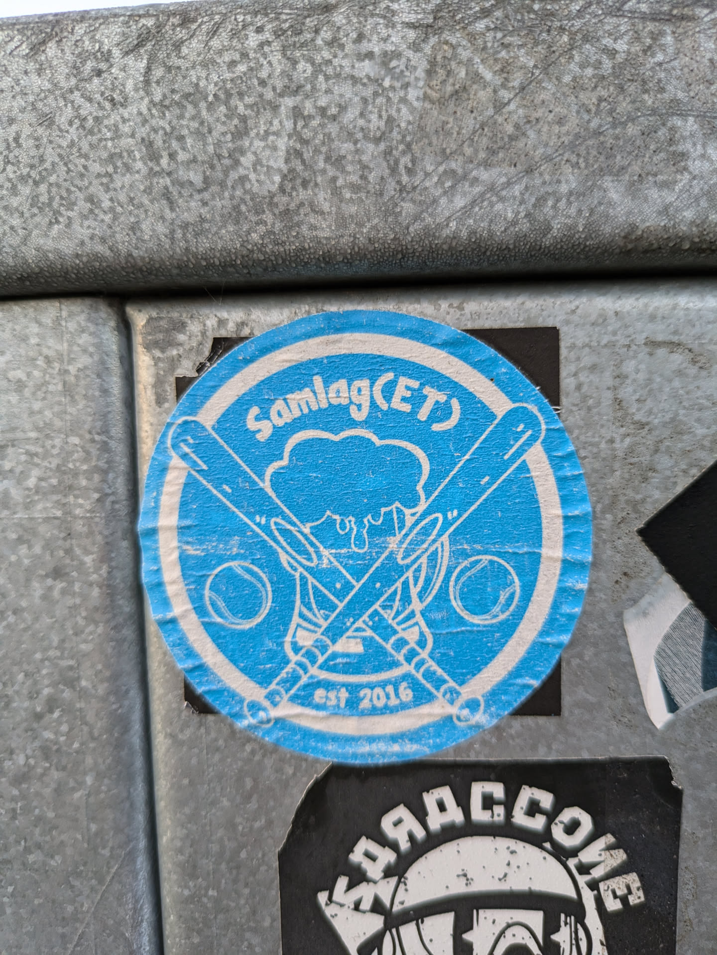 A blue circular sticker with a logo depicting crossed baseball bats behind a beer mug and two baseballs. The text 'Samlag(ET)' is written above the logo, with 'est 2016' below it.