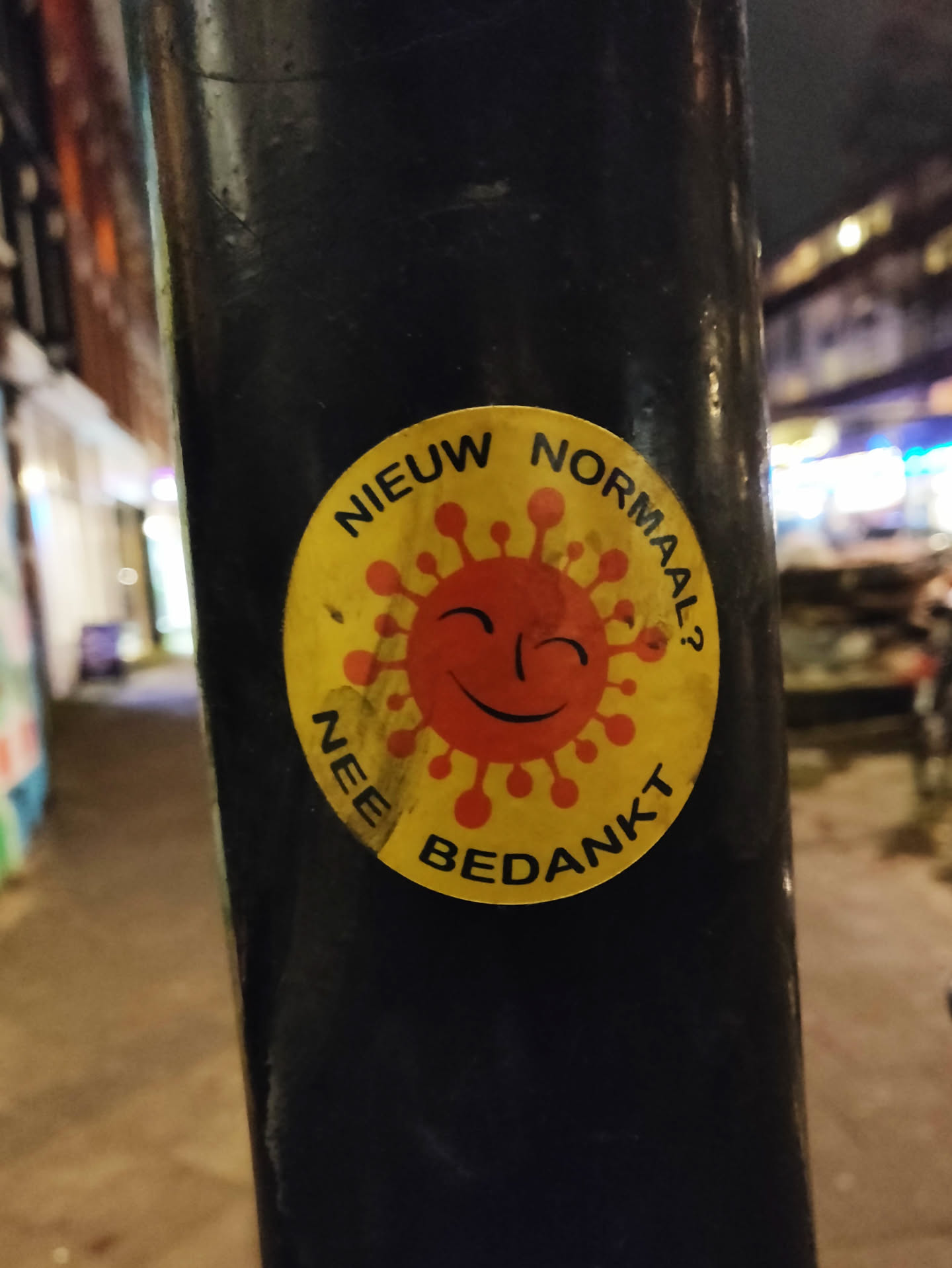 A round yellow sticker with a cartoon depiction of a virus with a smiling face.  The text around the image says "NIEUW NORMAAL? NEE BEDANKT" which translates from Dutch to "New Normal? No Thanks". The sticker is affixed to a dark-colored pole.