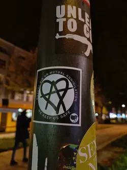 Black and white sticker with an anarchy symbol inside a heart. The text around says "Mutual Aid or Mutual Annihilation" and "We keep us safe".