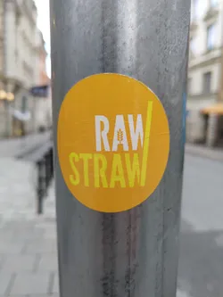 A round yellow sticker with the words RAW/STRAW in yellow text. A small wheat graphic is integrated into the 'A' of RAW.  The sticker is affixed to a grey metal pole on a city street.