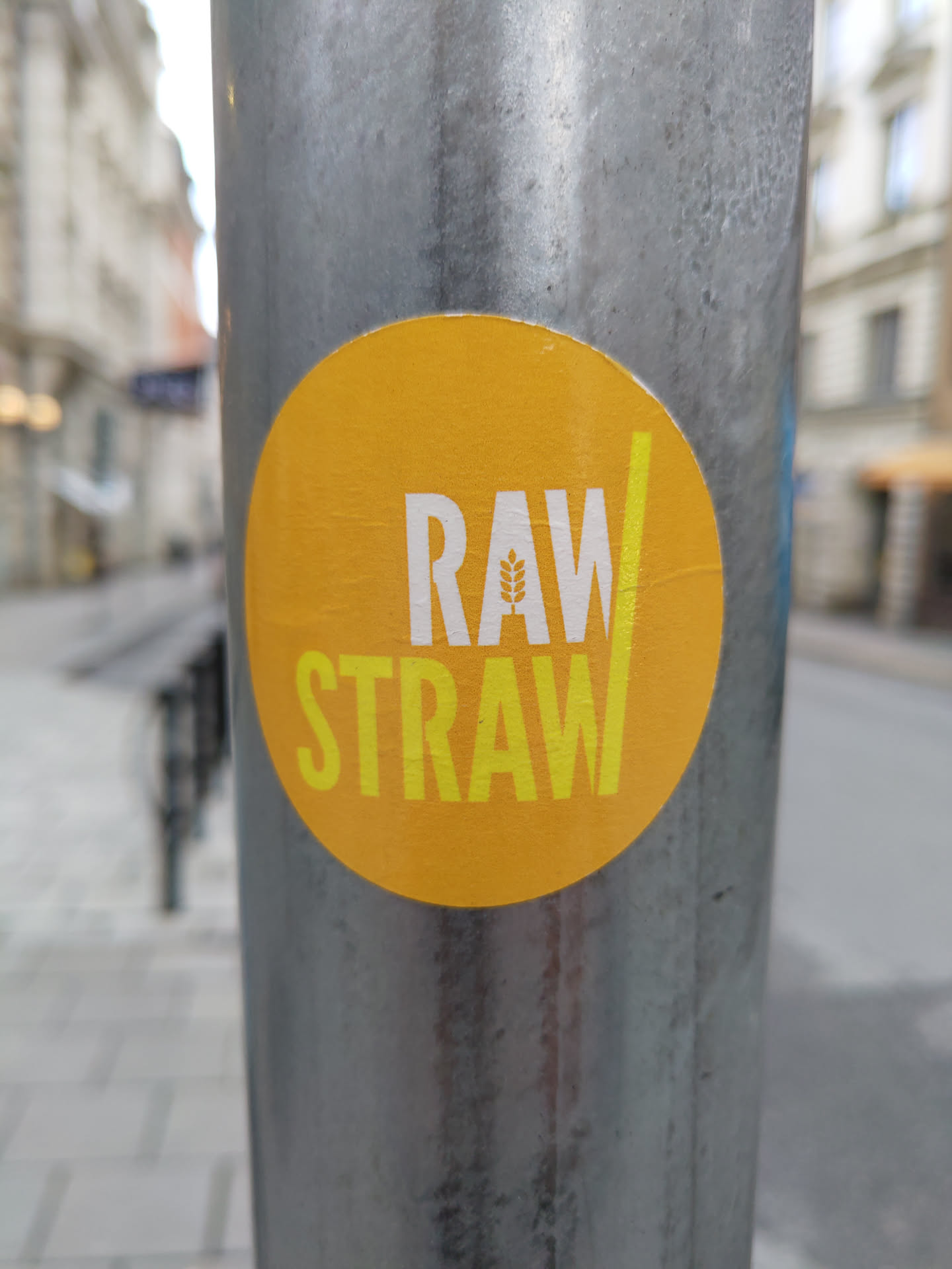 A round yellow sticker with the words RAW/STRAW in yellow text. A small wheat graphic is integrated into the 'A' of RAW.  The sticker is affixed to a grey metal pole on a city street.