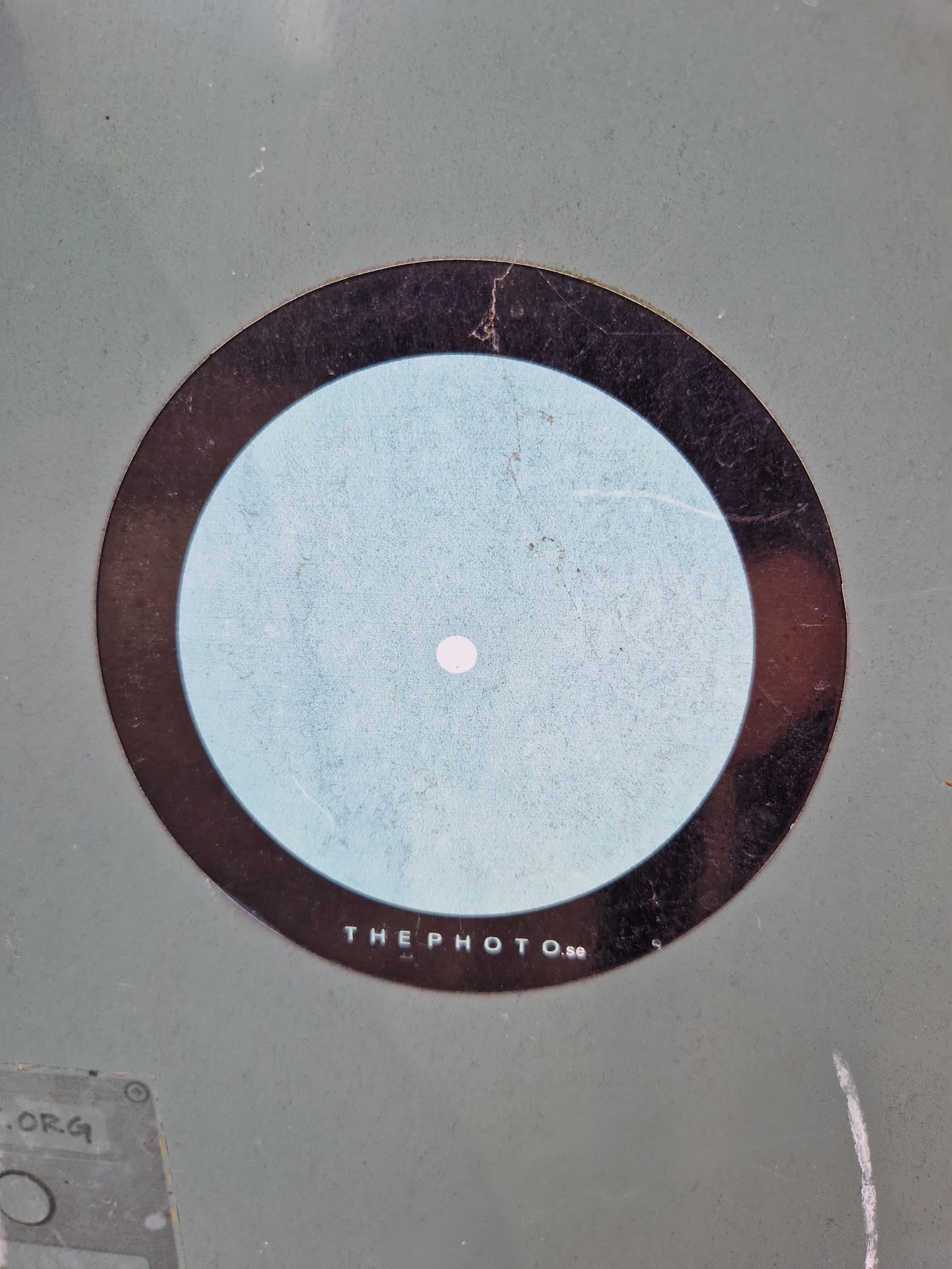 A round sticker featuring a concentric circle design. The outer ring is black, surrounding a larger light blue circle, which in turn contains a small white dot at its center.  The text "THE PHOTO..." is visible at the bottom of the sticker. The sticker appears to be affixed to a light greyish-green surface.