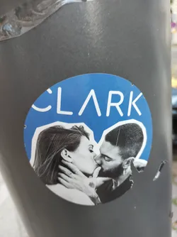 A round sticker features a black and white photograph of a couple kissing passionately against a bright blue background. The word 'CLARK' is prominently displayed in a modern, sans-serif font at the top of the sticker. The sticker shows some signs of wear and tear.