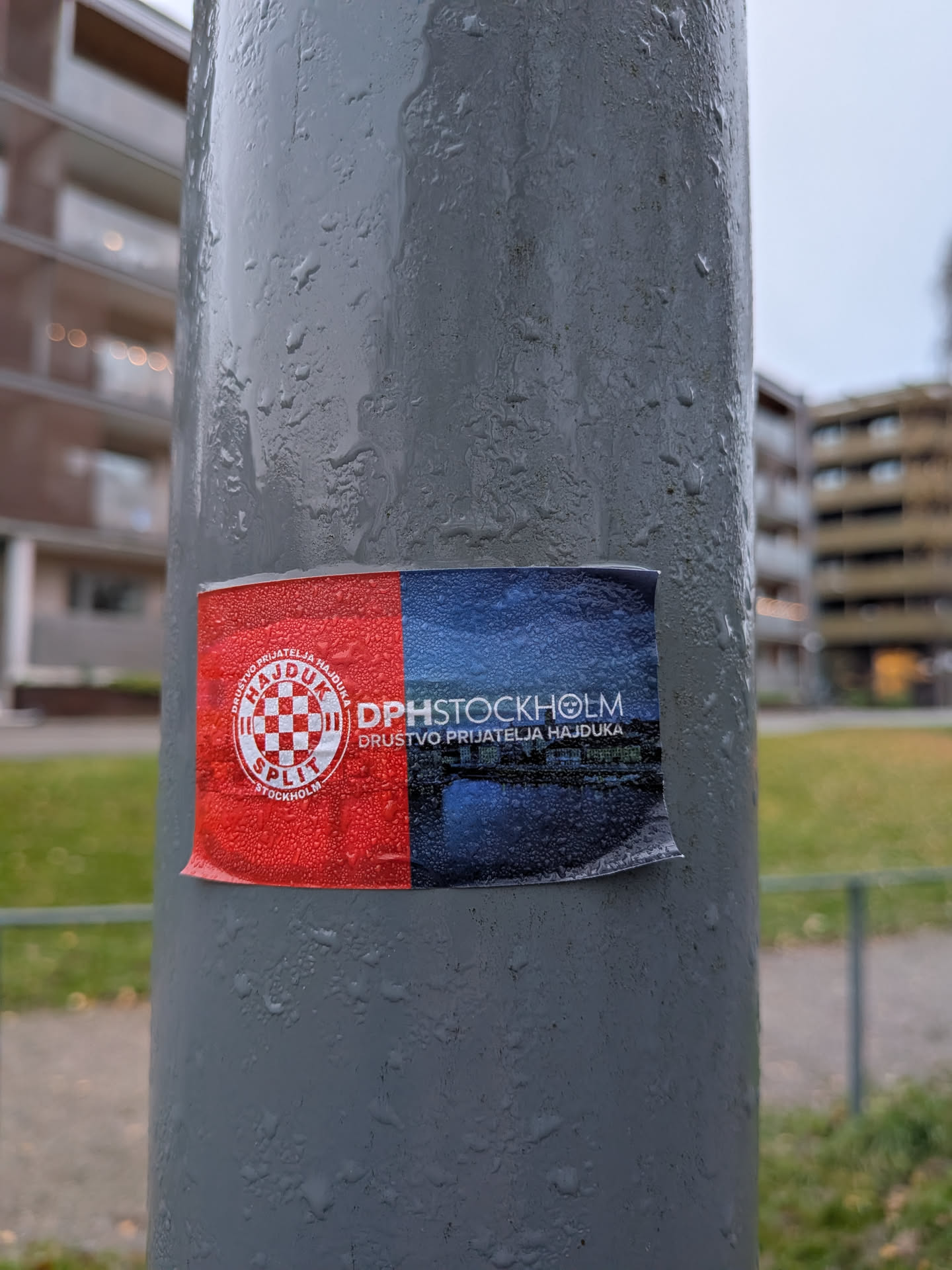 The sticker is horizontally divided into two halves. The left half is red and features a circular emblem with a checkered pattern in white and red, resembling a crest. Text surrounds the crest, including 'HAJDUK SPLIT' and 'STOCKHOLM'. The right half of the sticker is dark blue and depicts a nighttime cityscape with illuminated buildings reflected in water, likely representing Stockholm. The text 'DPHSTOCKHOLM' and 'DRUŠTVO PRIJATELJA HAJDUKA' are prominently displayed across this section. The sticker appears to be slightly creased and has water droplets on its surface.