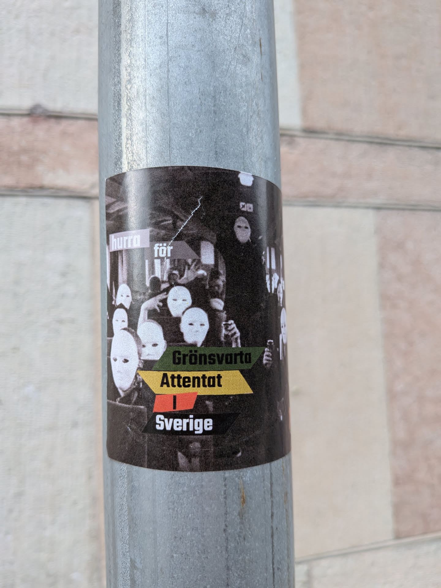 A rectangular sticker with rounded corners affixed to a metal pole. The background is a black-and-white photograph depicting a crowd of people wearing blank white masks. Overlaid on the image is text in various colored blocks: 'hurra' in a light pink box, 'för' in a grey box, 'Grönsvarta' on a green banner, 'Attentat' on a yellow banner, 'i' on a red banner, and 'Sverige' on a black banner.