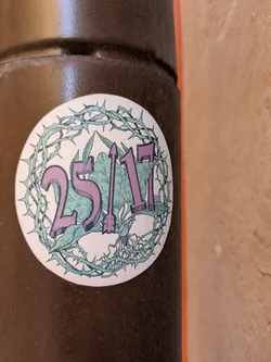 The sticker is circular and features the text "25/17" in purple, with an arrow replacing the slash. The background includes green foliage and a skull. A thorny wreath surrounds the design.