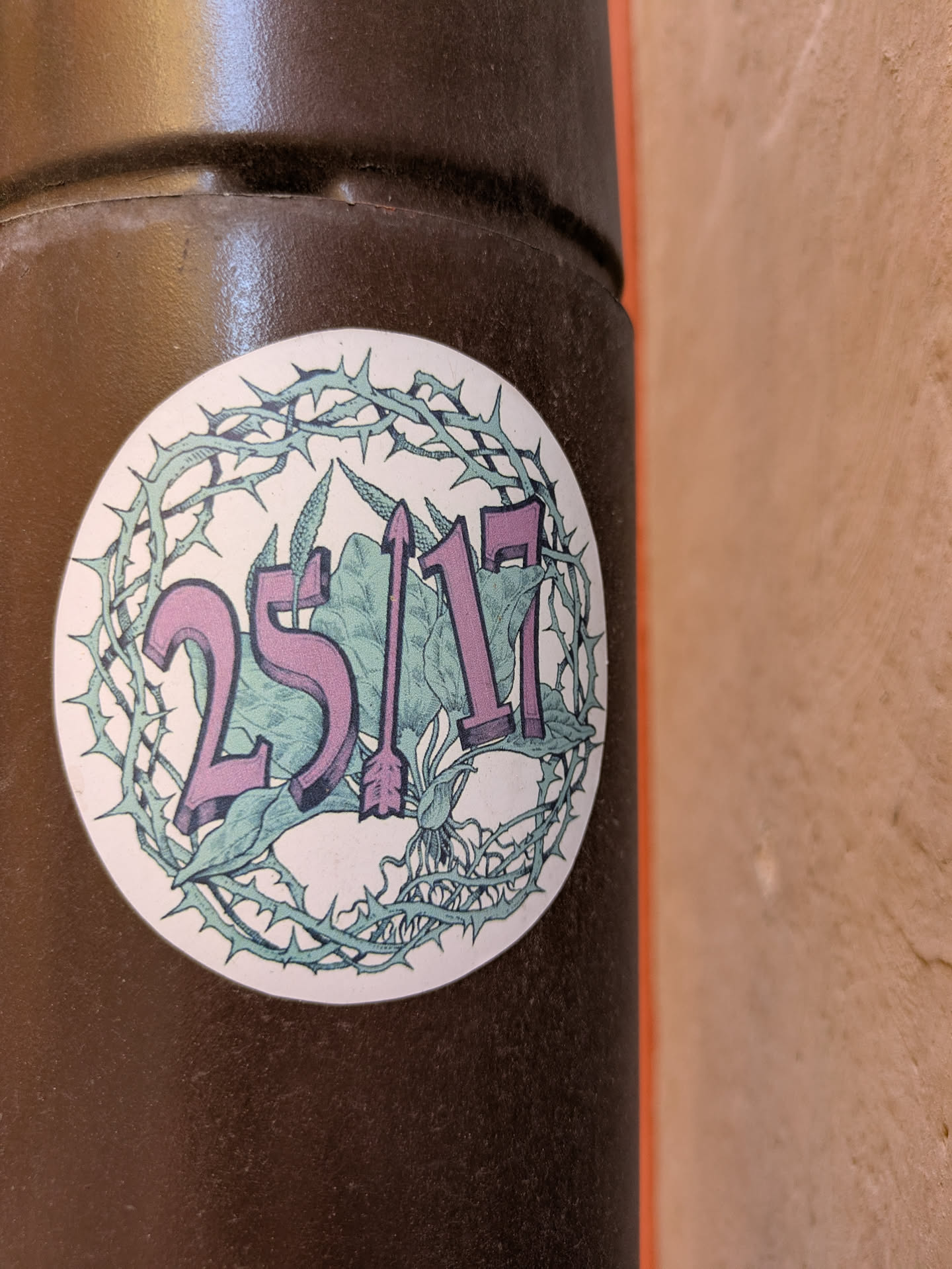 The sticker is circular and features the text "25/17" in purple, with an arrow replacing the slash. The background includes green foliage and a skull. A thorny wreath surrounds the design.