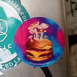 Round sticker featuring an illustration of a double cheeseburger topped with a cluster of mushrooms. The background is a tie-dye style in shades of pink, purple and blue.  The artist's Instagram handle is visible at the bottom.