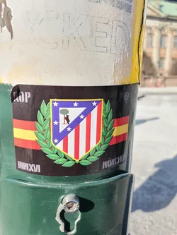 A sticker featuring the Atletico Madrid crest, surrounded by a laurel wreath, with MMXVI (2016) and MUNCHE written on it. The sticker is attached to a green pole.