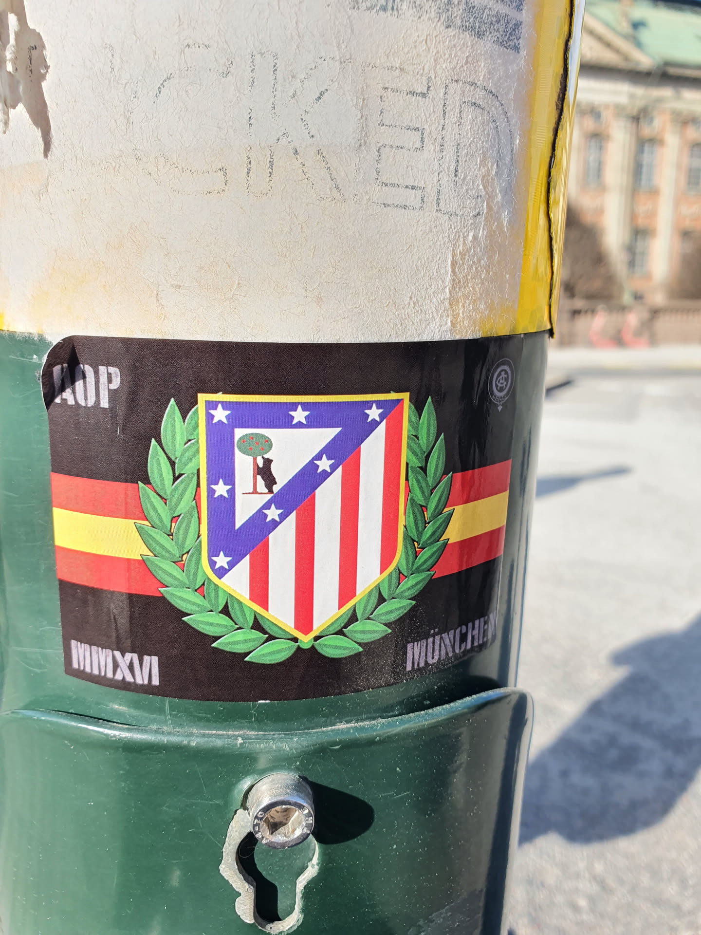 A sticker featuring the Atletico Madrid crest, surrounded by a laurel wreath, with MMXVI (2016) and MUNCHE written on it. The sticker is attached to a green pole.