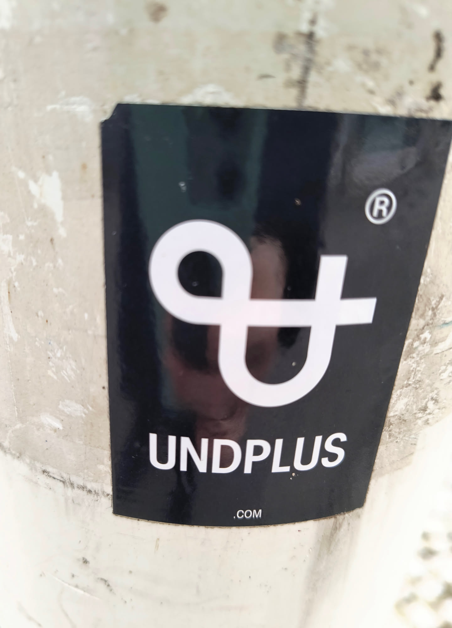 A black sticker with the logo of UNDPLUS on a white pole. The logo consists of two linked "U" shapes, one rotated 90 degrees.  Below the logo is the text "UNDPLUS .COM".